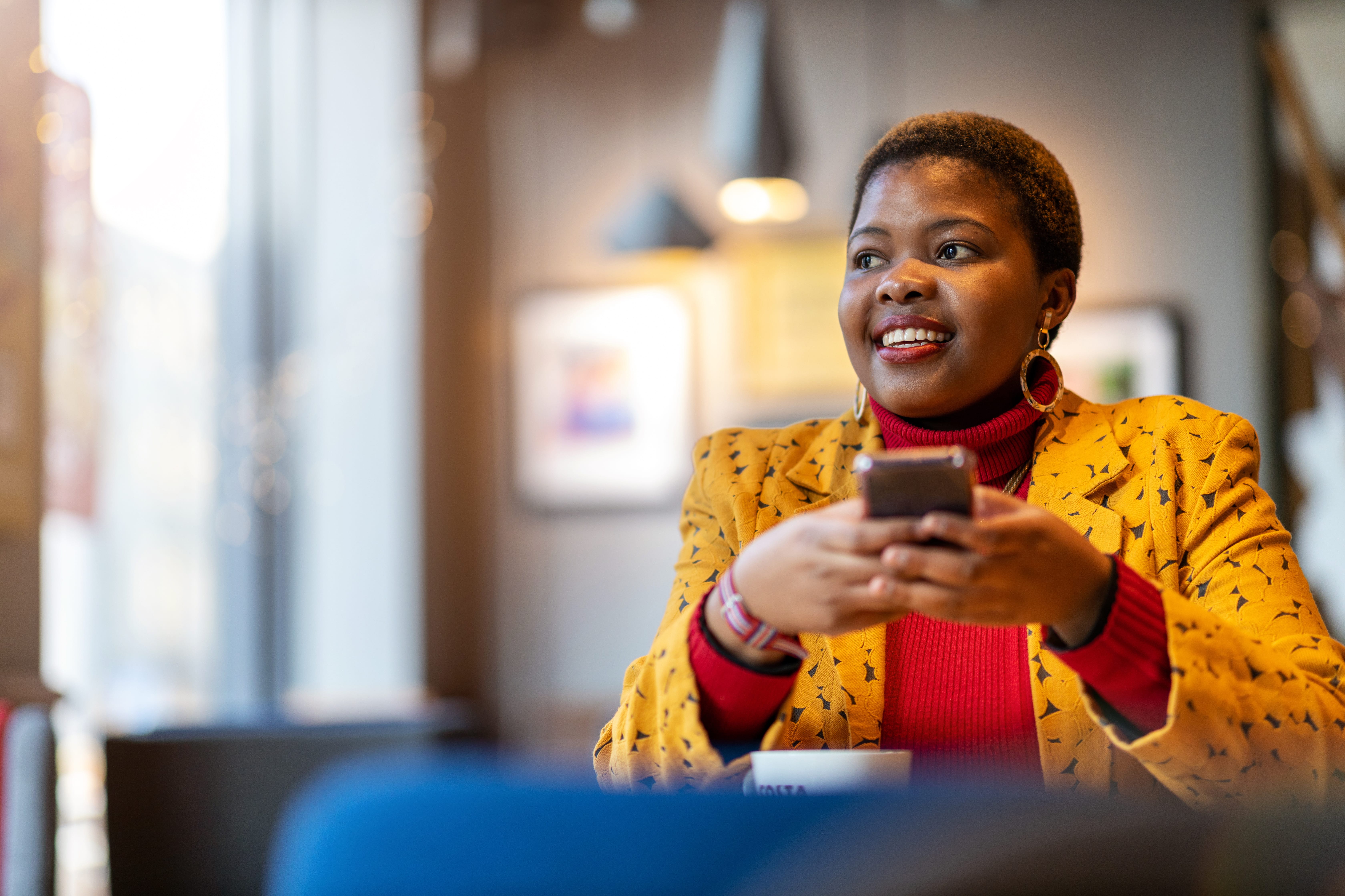 Young lady wearing a red turtle neck sweater and yellow suit jacket smiling as she holds her cell phone, while drinking a cup of coffee.