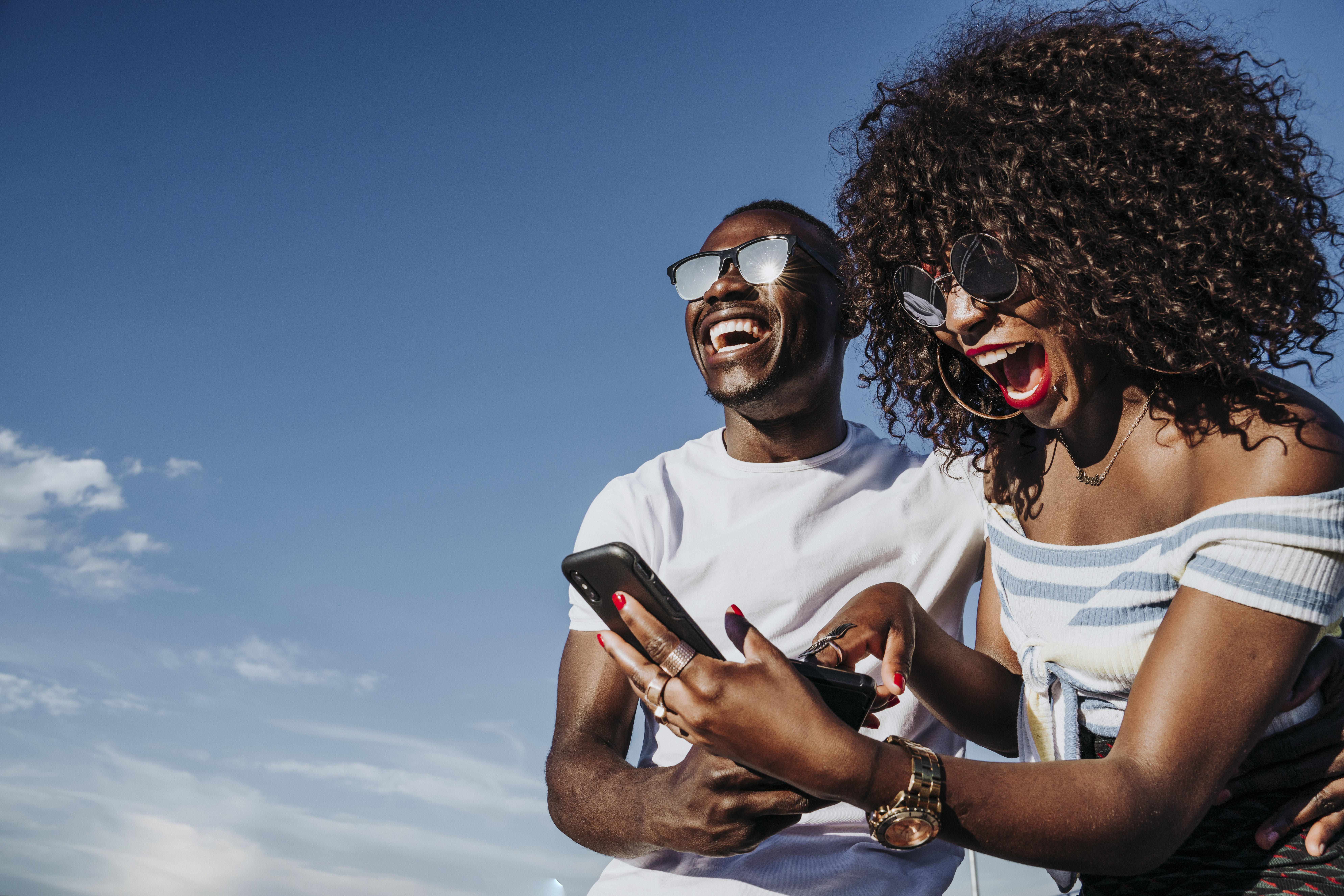 A young, African couple smiling and laughing on a bright, sunny day as the woman looks at her cell phone.