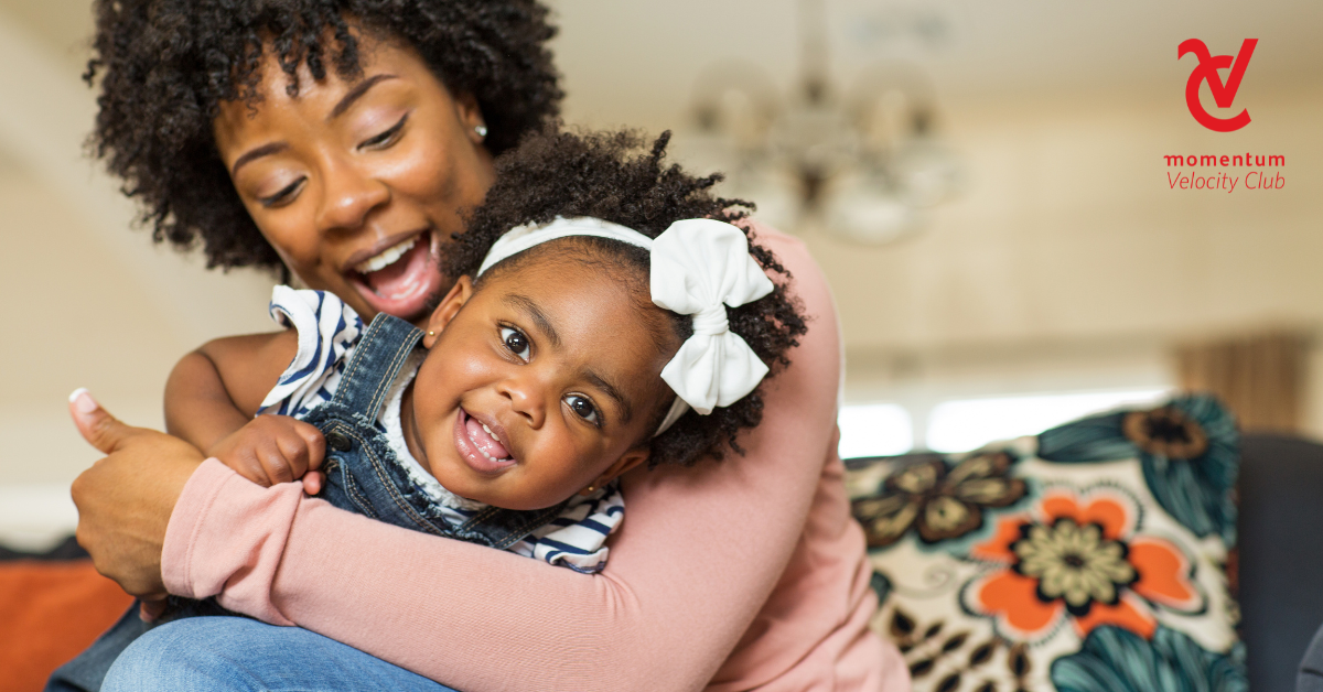A happy young mom hugs her young daughter while seated on a couch.