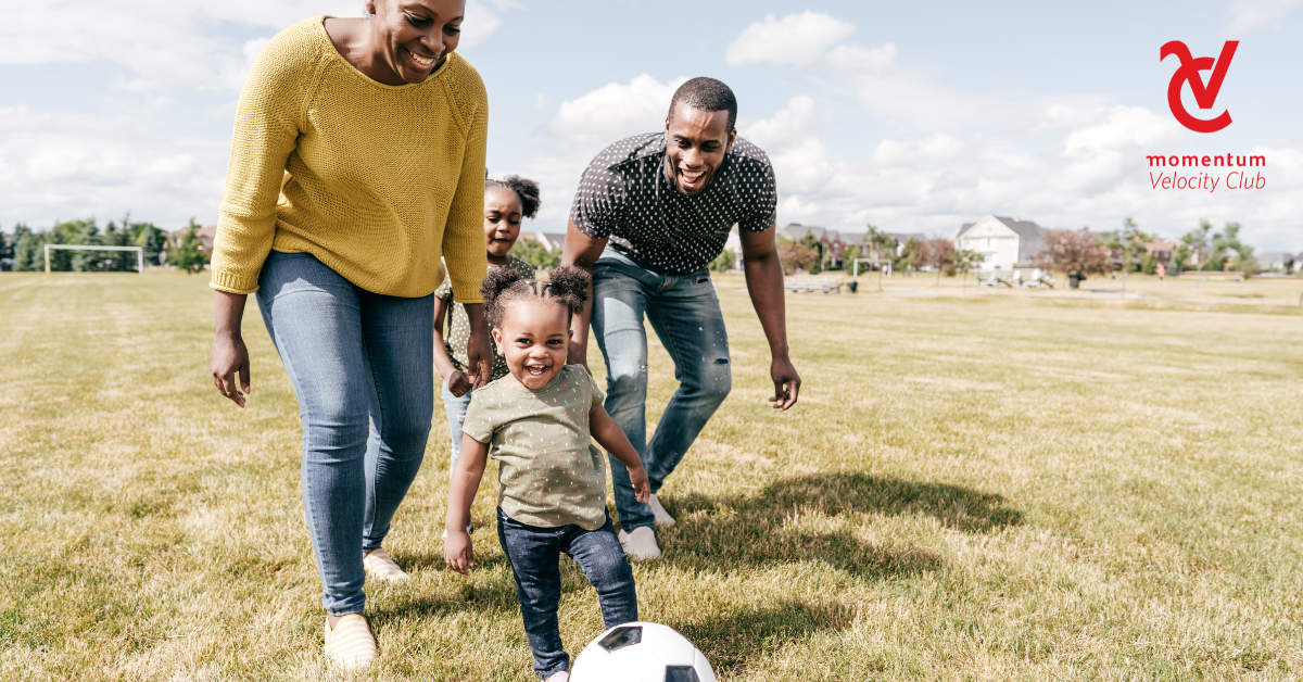 A mom and dad are having fun playing soccer in the park with their two young children.
