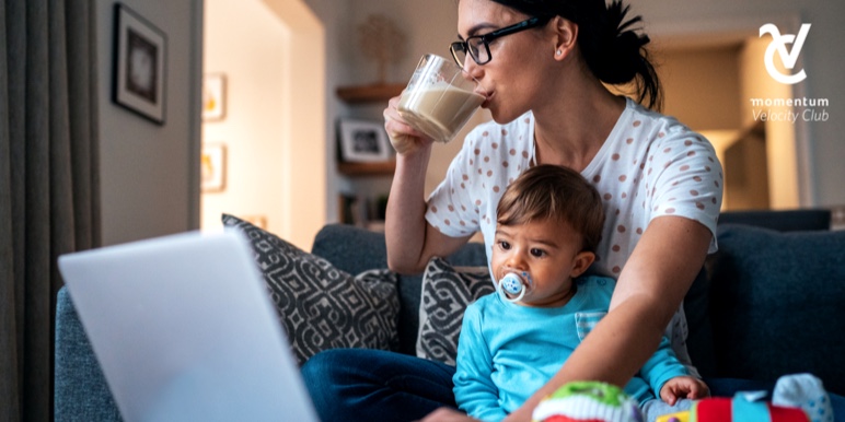 A young parent is sitting on a couch and working on her budget on her laptop. She’s drinking coffee while her young son sits next to her.