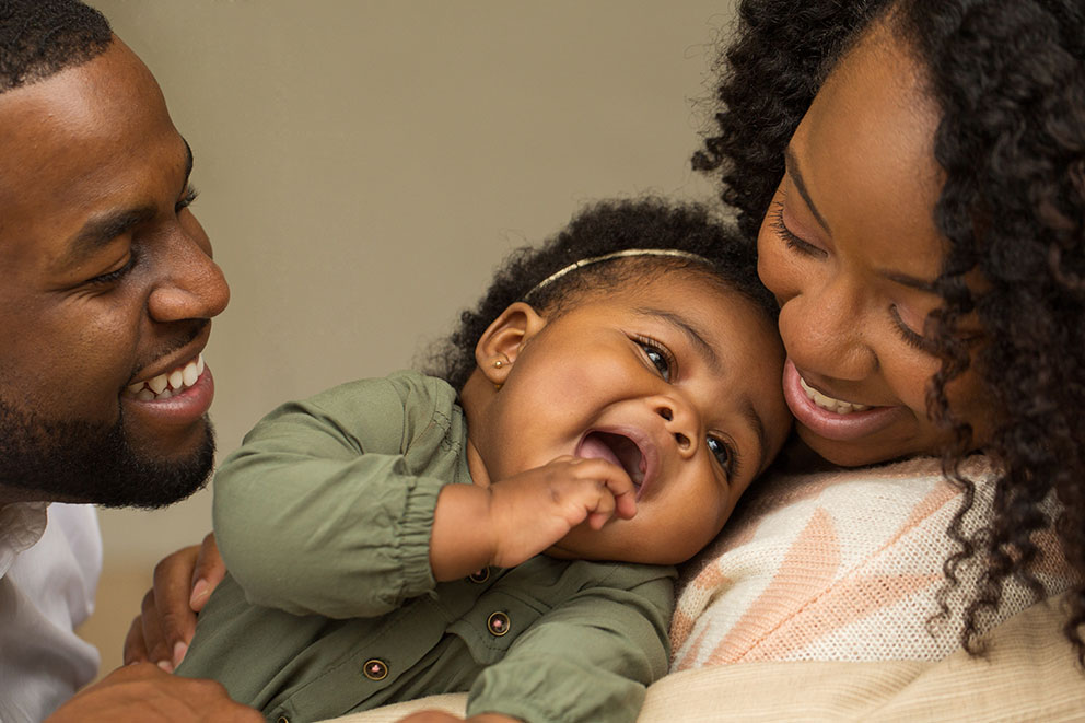 Young, African mom and dad smiling. The mom is holding a young toddler who is laughing.