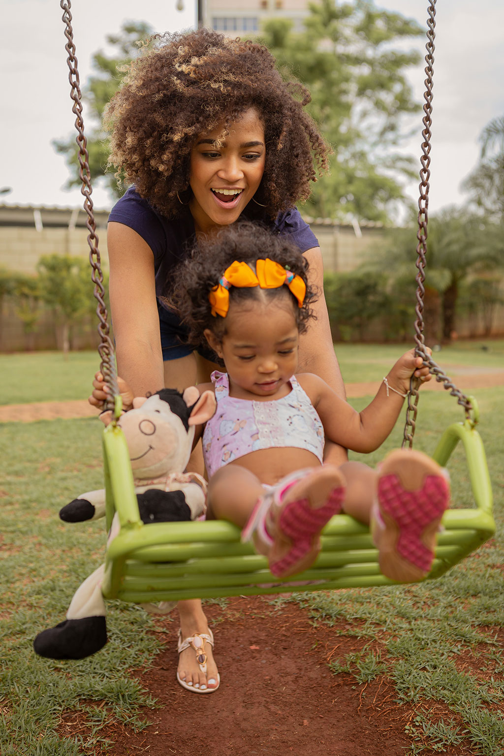A young mom smiling as she pushes her daughter and her cow plush toy on a swing.