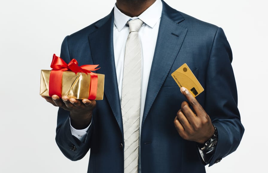 Man in a smartly dressed suit holds a Christmas gift in one hand and a credit card in the other.