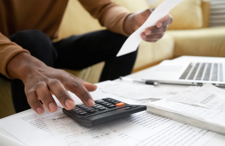 A man seated in front of a desk with lots of paperwork and his laptop, while he is working out his tax calculations on a calculator.