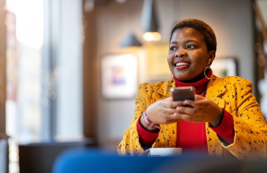 Young lady wearing a red turtle neck sweater and yellow suit jacket smiling as she holds her cell phone, while drinking a cup of coffee.