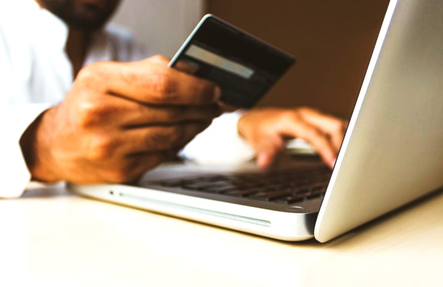 A man wearing a white shirt working on his laptop with one hand and holding his current account bank card in his other hand.