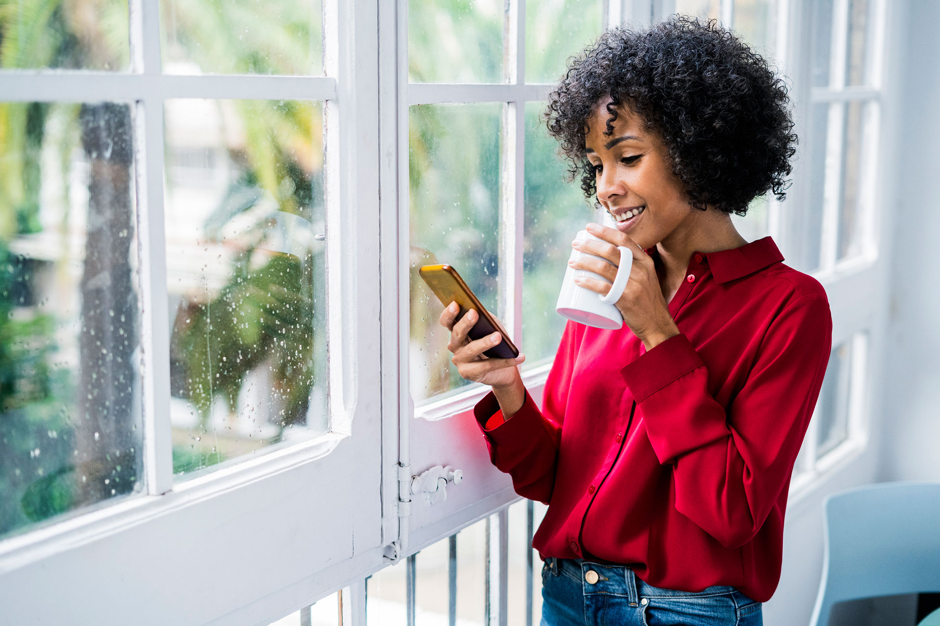 Young woman drinking a cup of coffee and holding her cell phone while standing in front of a window while it rains outside.