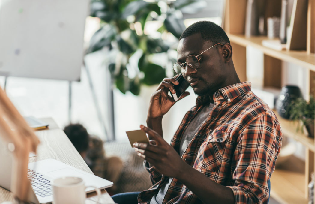 Man in casual shirt sitting next to his laptop holding phone with his card in the other hand.